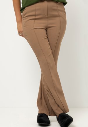 Pantalon brun extensible avec une coupe ajustée, présentant des détails de couture sur le devant et une texture lisse, associé à des chaussures à enfiler en fourrure noire.