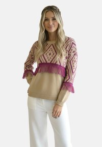 Sweter dziany z beżową bazą i różowym geometrycznym wzorem, z frędzlami przy dolnym brzegu i mankietach oraz prążkowanymi detalami przy dekolcie.