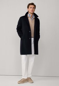 Cappotto di lana blu navy con colletto ampio, chiusura con bottoni, indossato sopra un maglione beige e una camicia azzurro chiaro, coordinato con pantaloni bianchi.