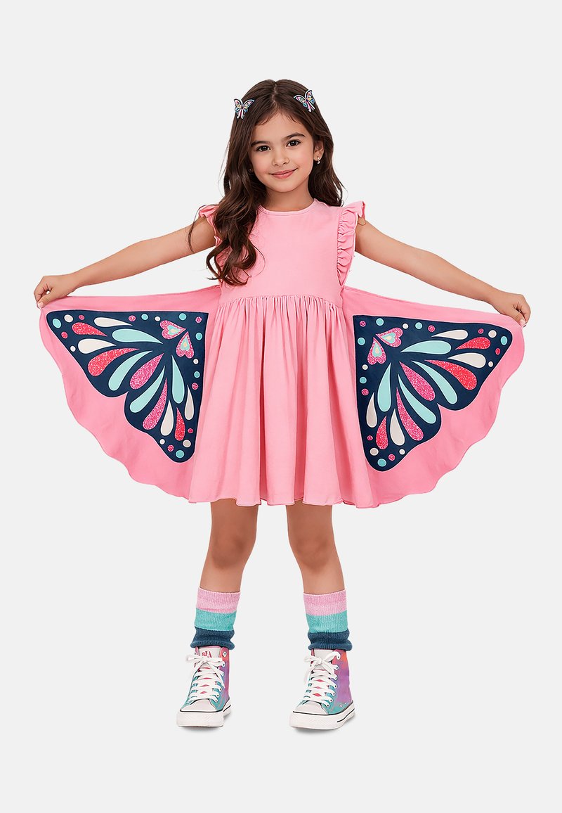 Ung pige i en pink kjole med sommerfuglevinge-mønstre, iført sommerfugle-hårclips, stripede sokker og farverige sneakers, spreder sine arme vidt.