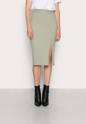 Miętowo zielona spódnica w prążki sięgająca do kolan z przednim bocznym rozcięciem, noszona z białą włożoną do środka bluzką i czarnymi botkami za kostkę.