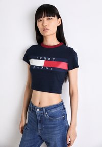 Tamsiai mėlyna trumpa marškinė su raudona ir balta horizontalia juostų dizainu bei "TOMMY JEANS" tekstu. Minkšta medvilnė su raudonu raišteliu.