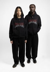 Black zip-up hoodies with red and white flame-like graphic details, paired with relaxed black pants and black shoes, on two figures.