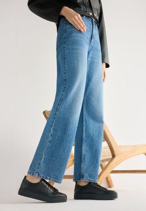 Osoba ubrana w szerokie niebieskie dżinsy, czarną skórzaną kurtkę i czarne trampki stoi obok drewnianego plecionego krzesła na jasnej podłodze.