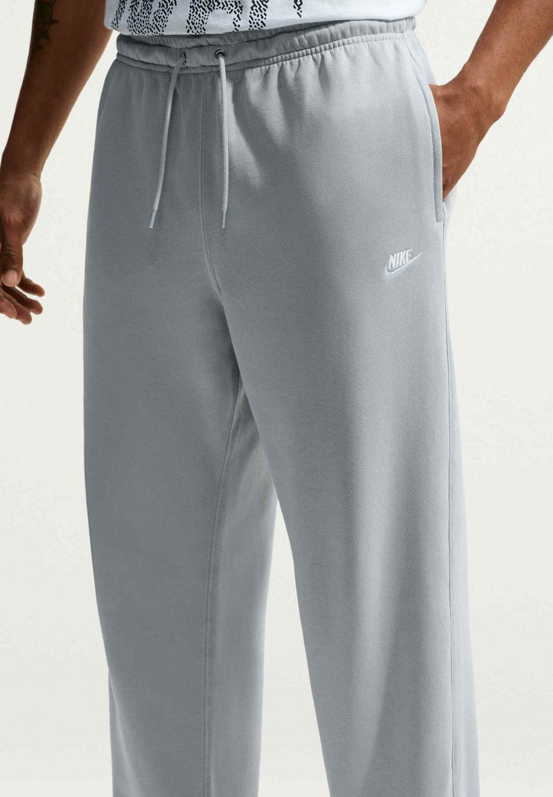 Afslappede lysegrå Nike joggingbukser med snøre i taljen, sidelommer og hvidt Nike-logo på venstre lår, båret af en person.