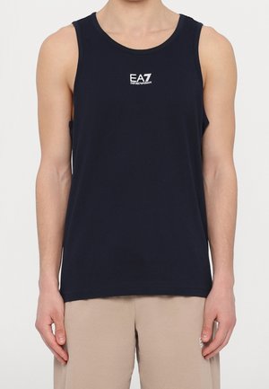 Mężczyzna ubrany w granatową, bezrękawnikową koszulkę z białym logo EA7 Emporio Armani na piersi oraz beżowe szorty.