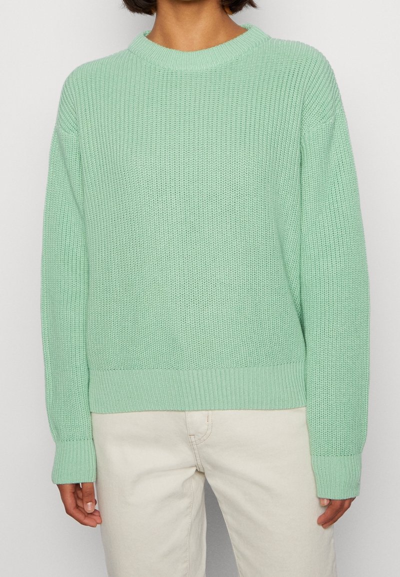 Maglione lavorato a maglia verde chiaro con texture a coste, collo rotondo e maniche a cuffia, abbinato a pantaloni di colore chiaro.
