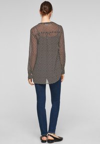 Blusa nera trasparente con un motivo geometrico, maniche lunghe, orlo arrotondato, abbinata a jeans skinny blu scuro e scarpe flat nere.