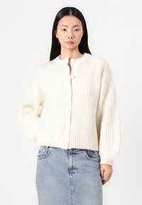 Krémový pletený cardigan s okrúhlym výstrihom, rebrovanou textúrou a veľkými gombíkmi. Kombinovaný s svetlomodrou džínsovou sukňou.