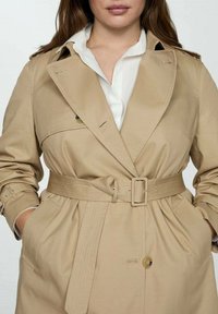 Manteau beige avec taille ceinturée, grandes poches latérales et col à revers classique. Fabriqué en tissu lisse avec fermeture à boutons.