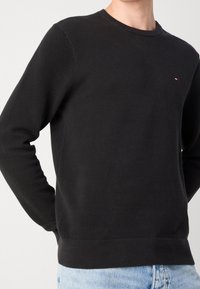 Schwarzer Sweatshirt mit texturiertem Stoff, rundem Halsausschnitt und langen Ärmeln. Beinhaltet ein kleines gesticktes Logo in Rot und Weiß auf der Brust.