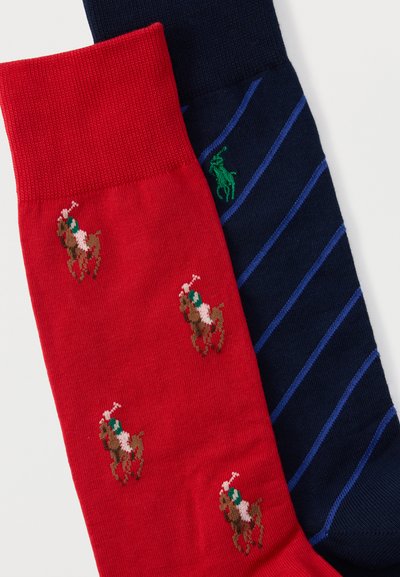 Κόκκινη κάλτσα με καφέ σχέδιο αλόγου και αναβάτη δίπλα σε μπλε σκούρα κάλτσα με διαγώνιες μπλε ρίγες και πράσινο λογότυπο αλόγου και αναβάτη.