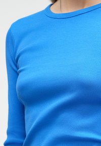 Κοντινή λήψη ενός ατόμου που φοράει ένα εφαρμοστό ριμπ πλεκτό μπλε πουλόβερ με μακριά μανίκια, εμφανίζοντας το πάνω μέρος του κορμού και τη γραμμή του λαιμού.