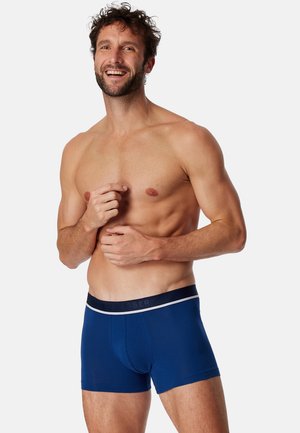 Sous-vêtement type trunk bleu marine en tissu doux, avec une coupe ajustée, une ceinture contrastante portant le nom de la marque, et des jambes courtes.