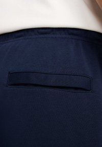 Tumši zils audums ar gludu faktūru. Aizmugurē ir horizontāla atvērta kabata. Šuvju detaļas ir redzamas gar malām.