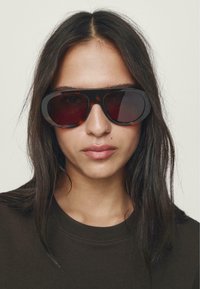 Runde solbriller med mørke linser og en tyk skildpaddefarvet ramme. Modellen har en mørk skjorte på og langt, glat hår.