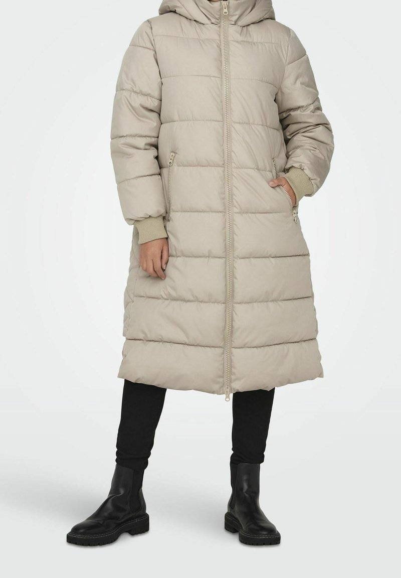 Beige donsjack met capuchon, met doorgestikte delen, een volledige ritssluiting en zijzakken. Gecombineerd met zwarte leggings en enkellaarsjes met hak.