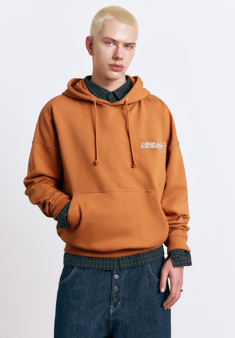 Jeune personne aux cheveux blonds courts portant un sweat à capuche orange par-dessus une chemise à carreaux vert foncé et un jean bleu foncé, debout devant un fond uni.