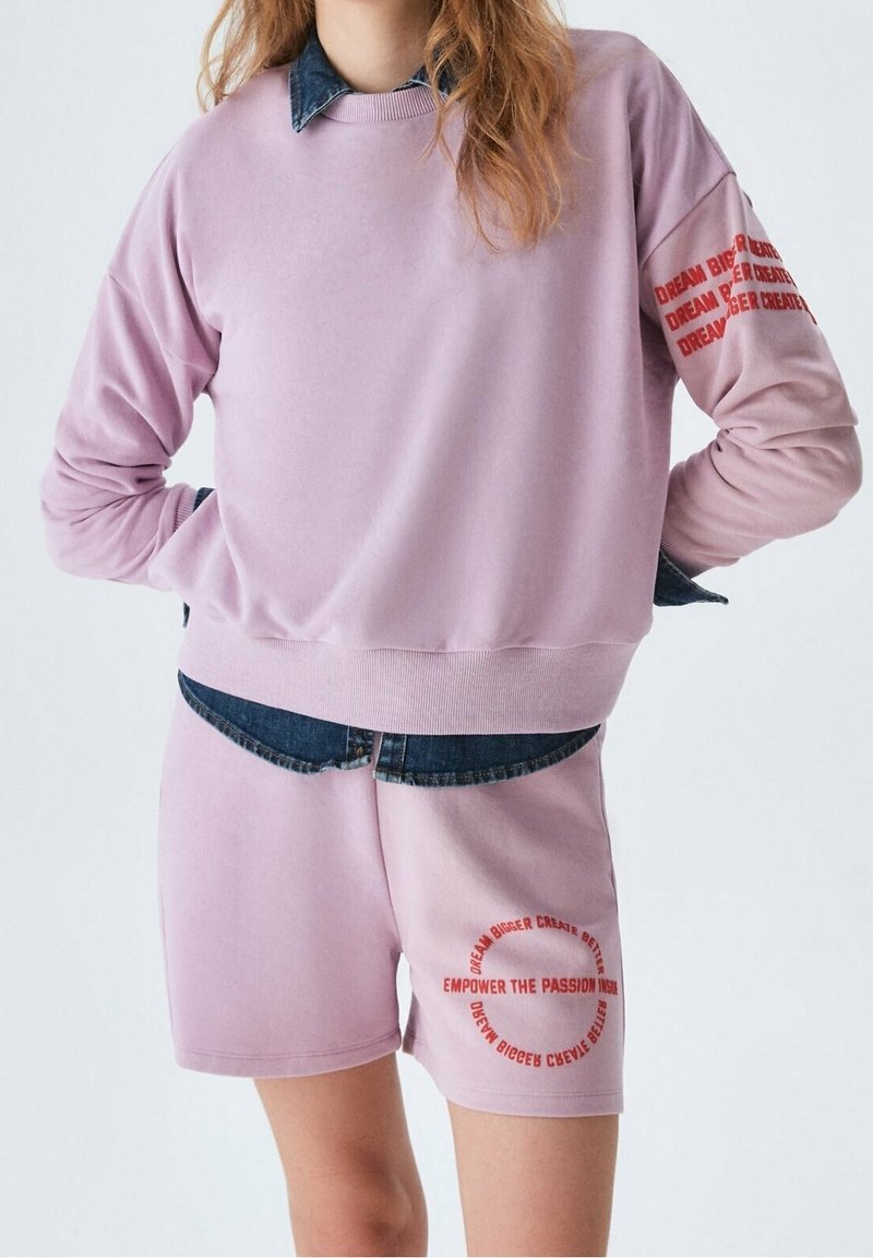 Person som bär en ljuslila sweatshirt och shorts med röd text, i lager över en mörkblå denimskjorta, stående mot en neutral bakgrund.