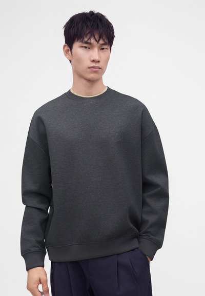 Jeune homme aux cheveux noirs courts, portant un sweat-shirt gris foncé et un pantalon bleu marine, debout sur un fond blanc uni.