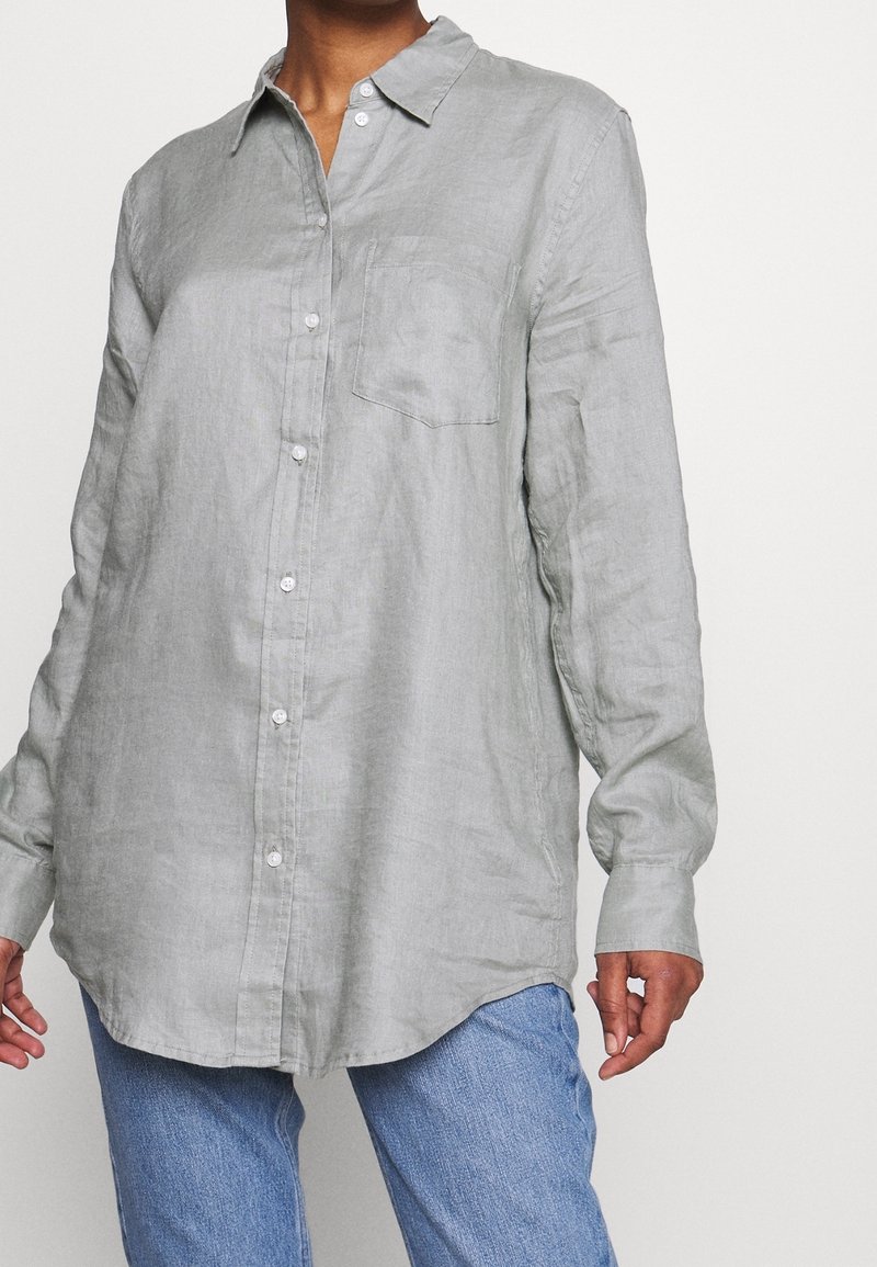 Camisa de botones de manga larga en gris claro, hecha de lino, con cuello, detalle de bolsillo y dobladillo redondeado. Usada con jeans azules.