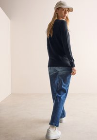 Svart långärmad skjorta i kombination med ljusblå denimjeans. Vita sneakers och en beige keps fulländar den avslappnade outfiten. Neutral bakgrund.