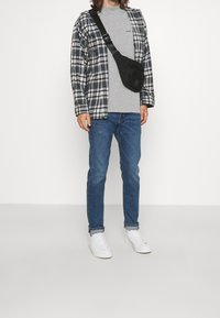 Rutig svart och vit jacka över en grå t-shirt, blå jeans och vita sneakers. Axelväska placerad över axeln.