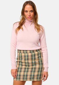 Femme portant un pull à col roulé rose clair et une mini-jupe en plaid beige, se tenant avec une expression neutre devant un fond uni.