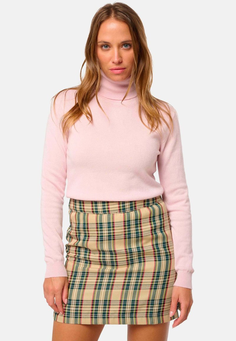 Femme portant un pull à col roulé rose clair et une mini-jupe en plaid beige, se tenant avec une expression neutre devant un fond uni.
