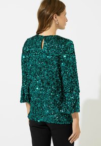Top verde con paillettes, caratterizzato da una scollatura rotonda, maniche a tre quarti e un dettaglio a chiave sulla schiena. La superficie è decorata con paillettes lucide e testurizzate.