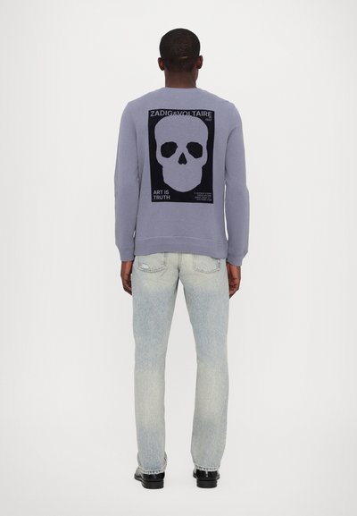 Sweatshirt violet avec un graphique de crâne noir au dos, comportant du texte. Porté avec un jean bleu clair déchiré et des chaussures noires.