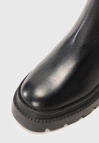 Close-up van een zwarte leren laars met een geribbelde dikke rubberen zool en een ronde teen op een witte achtergrond.
