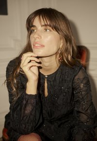 Rochie neagră brodată, cu mâneci lungi și bufante, având detaliu de gât cu deschidere în formă de cheie. Modelul poartă bijuterii din aur și pozează cu mâna aproape de față.