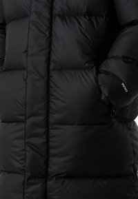 Mustast teksast puffer-jakk, millel on tikitud muster, esil lukk, ümarad varrukaääred ja väike logo varrukal.