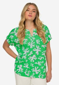 Top verde a maniche corte con scollatura a V, decorato con un motivo astratto bianco che ricorda forme di corallo. Tessuto leggero e vestibilità rilassata.