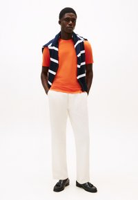 Πορτοκαλί μπλουζάκι με στρογγυλή λαιμόκοψη, λευκό παντελόνι, μπλε πουλόβερ με ρίγες περασμένο στους ώμους, μαύρα μοκασίνια. Απλή, καθημερινή εμφάνιση.