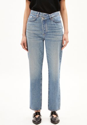 Person trägt hellblaue Jeans mit geradem Bein, asymmetrischem Taillenknopf und schwarzen Schnallenschuhen, steht vor weißem Hintergrund.