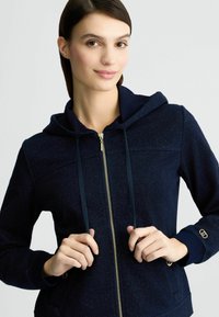 Felpa con zip di colore navy, dotata di cappuccio e lacci, caratterizzata da una zip metallica e un tessuto testurizzato con un leggero luccichio.
