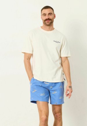 Mann mit Schnurrbart, der ein weißes T-Shirt und blaue gemusterte Shorts trägt, steht mit einer Hand in der Tasche vor einem schlichten hellen Hintergrund.