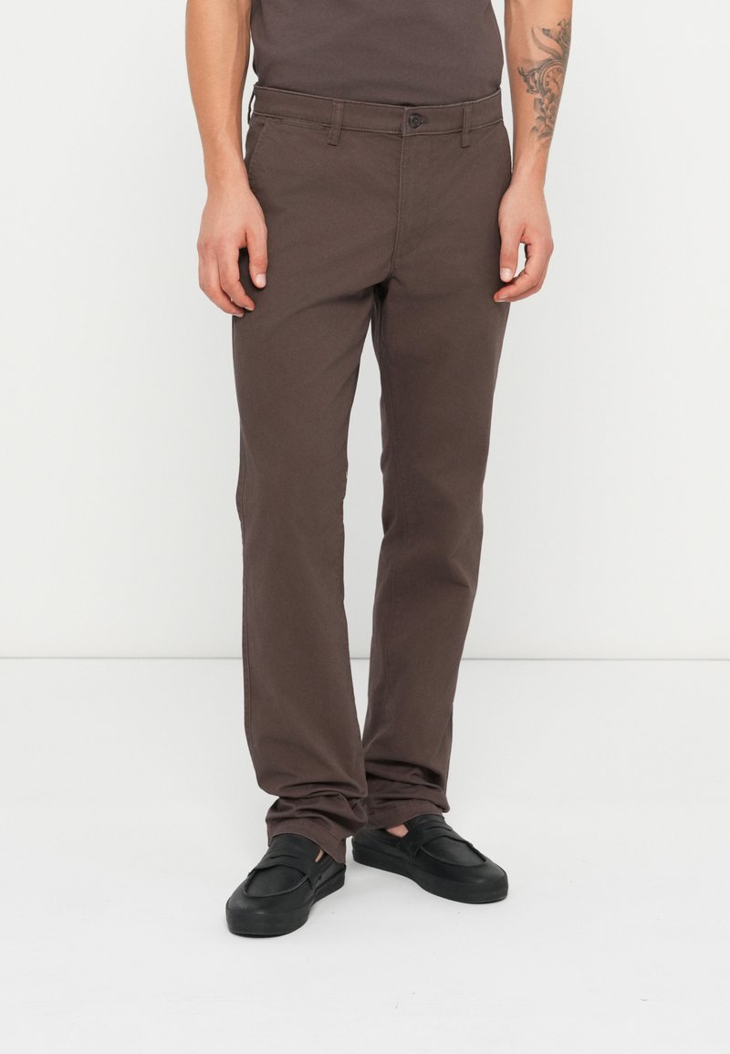 Brune bomulds chinos med lige snit, flad front og sidelommer; parret med sorte slip-on sko. Glat tekstur og minimalistisk design.
