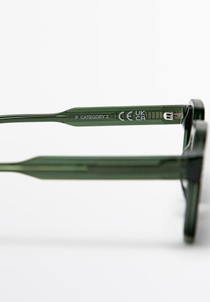 Close-up af grøn brillearm med teksten "P KATEGORI 3" samt CE- og UKCA-certificeringsmærker på hvid baggrund.