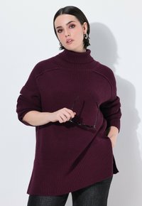 Burgundowy sweter prążkowany z wysokim kołnierzem, opadającymi ramionami i bocznymi rozcięciami. Model trzyma czarne okulary przeciwsłoneczne, stojąc na białym tle.