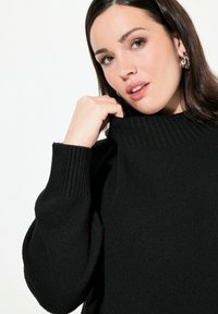 Czarny sweter dzianinowy z wysokim prążkowanym kołnierzem, z szerokimi rękawami i miękką teksturą. Model delikatnie trzyma kołnierz, pokazując jego design.