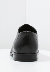 Chaussure habillée en cuir noir avec une finition lisse, bout arrondi, minimalisme dans les coutures, et une semelle en caoutchouc plate.