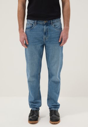 Hombre vistiendo jeans de mezclilla azul claro, camisa negra y zapatos negros con cordones, de pie sobre fondo blanco.