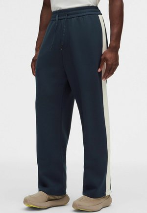 Mand iført marineblå joggingbukser med hvide sidelister og beige slip-on sko, stående mod en enkel hvid baggrund.