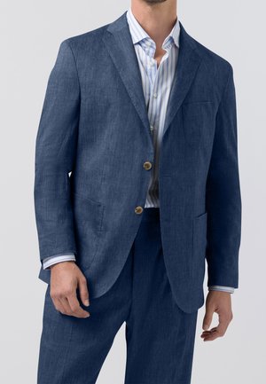 Mand iført en blå linnedragt med to knapper og en blå- og hvidstribet skjorte, stående foran en ensfarvet baggrund.