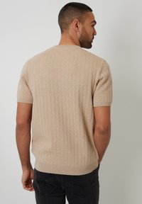 Beige gestrickter Pullover mit kurzen Ärmeln, strukturiertem Muster sowie geripptem Ausschnitt und Saum. Getragen mit dunklen Jeans, von hinten sichtbar.