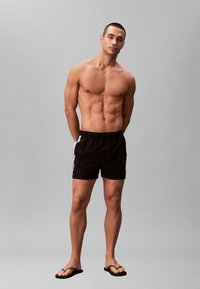 Man zonder shirt staand, gekleed in zwarte zwemboxers met zijstrepen en bruine slippers, tegen een effen grijze achtergrond.