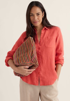 Kobieta z długimi ciemnymi włosami, ubrana w koralową koszulę zapinaną na guziki i beżowe spodnie, trzymająca pod pachą duży, wielokolorowy, pasiasty materiałowy kopertówkę.